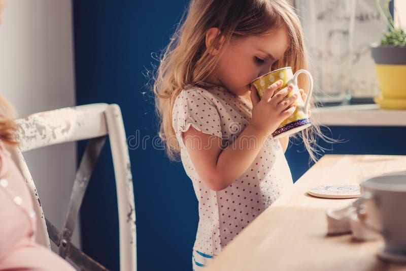 Cute baby girl drinking tea for breakfast in sunny kitchen