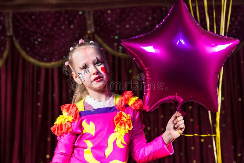 Cute blond girl wearing festive pink dress and clown make up while holding a star-shaped balloon, on a stage. Cute blond girl wearing festive pink dress and clown make up while holding a star-shaped balloon, on a stage