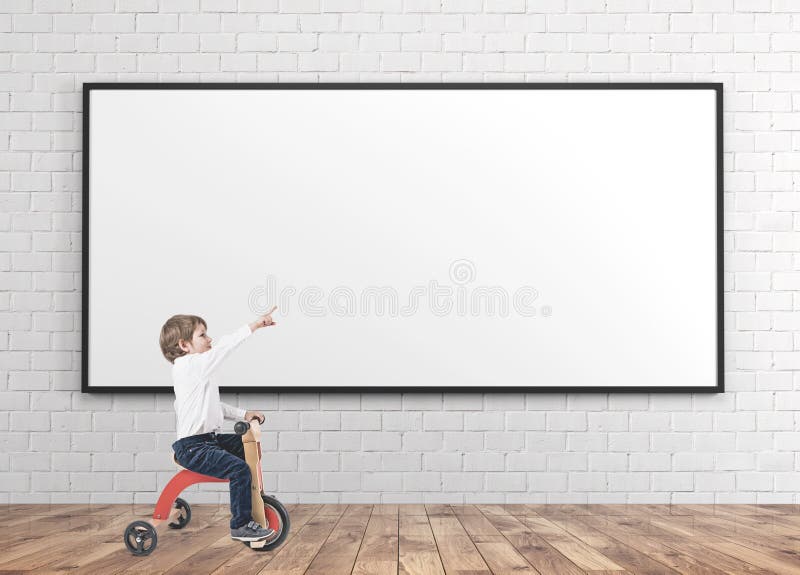 Cute little boy in a white shirt and dark blue jeans is riding a tricycle and showing with his finger. A wooden floor room with concrete walls. A blank whiteboard. Mock up. Cute little boy in a white shirt and dark blue jeans is riding a tricycle and showing with his finger. A wooden floor room with concrete walls. A blank whiteboard. Mock up