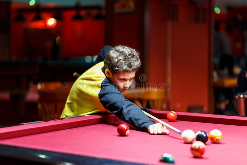 Cute boy in yellow t shirt plays billiard or pool in club. Young Kid learns to play snooker. Boy with billiard cue