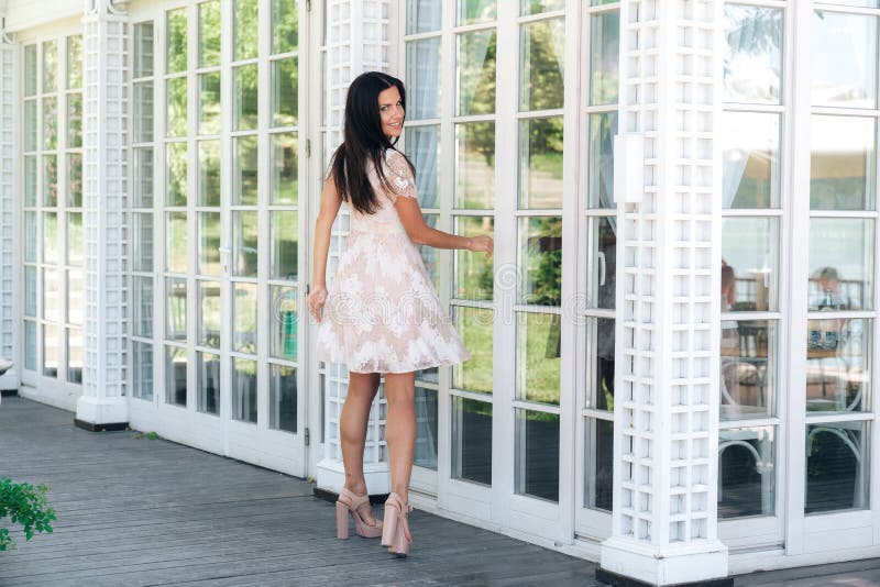 Cute brunette with beautiful long legs posing in beige colour dress outside near a wooden and glass wall stock photo Cute brunette with beautiful long legs posing in beige colour dress outside near a wooden and glass wall stock photo