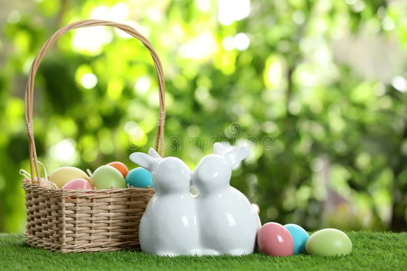 Cute ceramic Easter bunnies with wicker basket and dyed eggs on green grass against blurred background stock photography Cute ceramic Easter bunnies with wicker basket and dyed eggs on green grass against blurred background stock photography