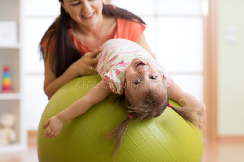 Cute child girl stretching on pilates fitness ball with mom in gym