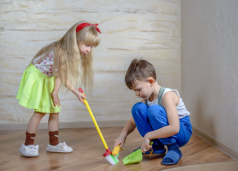 Cute little boy in blue pants, slippers and suspenders using toy broom and dustpan with girl in green skirt on hardwood floor. Cute little boy in blue pants, slippers and suspenders using toy broom and dustpan with girl in green skirt on hardwood floor