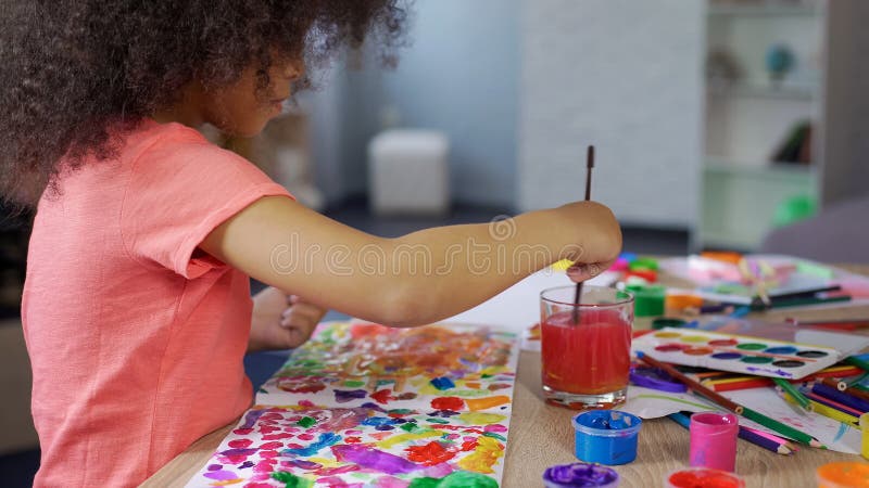 Cute curly African American girl putting paintbrush into water, leisure time royalty free stock photo Cute curly African American girl putting paintbrush into water, leisure time royalty free stock photo