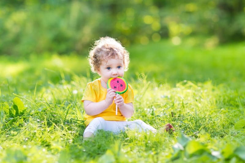 Cute curly baby girl eating watermelon candy in sunny park