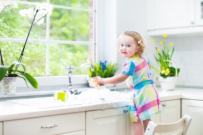 Cute curly toddler girl in colorful dress washing dishes