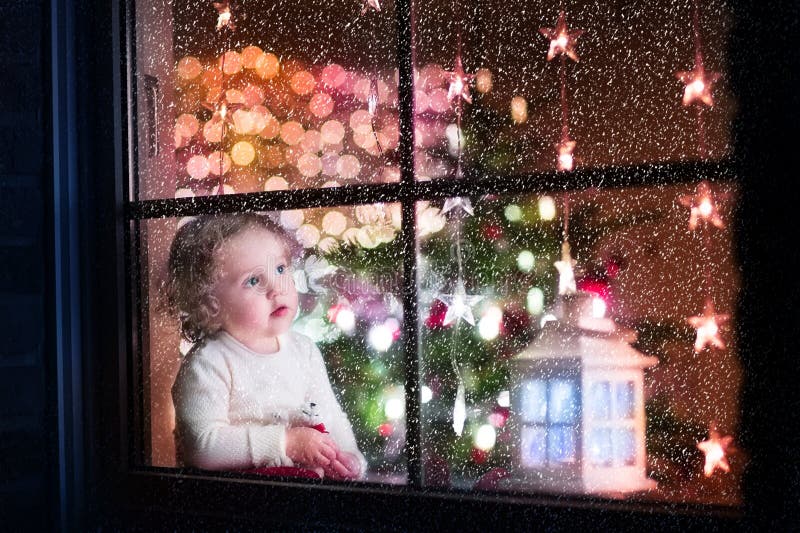 Cute curly toddler girl sitting with a toy bear at home during Christmas time, preparing to celebrate Xmas Eve, view through a window from outside into a decorated dining room with tree and lights