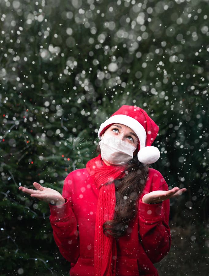 Cute European young woman in red Santa hat and protective mask from coronavirus sits next to the Christmas tree and catches white