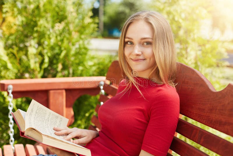 Cute female with freckled face, blonde hair, green charming eyes dressed in red clothes sitting at wooden bench in park reading bo