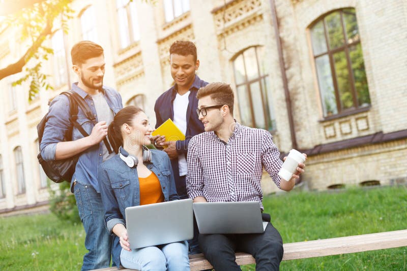 Happy young students are talking and smiling. Man and women are sitting on bench and holding laptops. Two guys are standing near them with joy. Happy young students are talking and smiling. Man and women are sitting on bench and holding laptops. Two guys are standing near them with joy