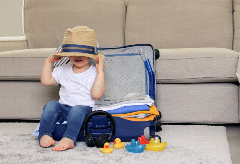 Cute funny little baby boy siiting in blue suitcase with hat on his eyes, packed for vacation full of clothes ready for traveling.