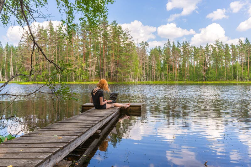 Cute ginger girl sitting on wooden planked footway and working with laptop in summer day against beautiful landscape of northern