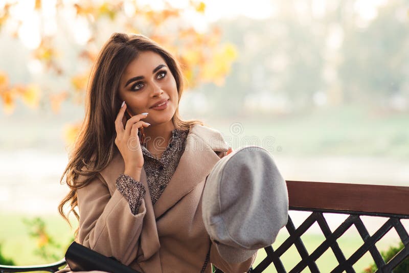 Cute girl flirts while talking on the phone with her boyfriend. Pretty woman sitting on the bench in autumn park