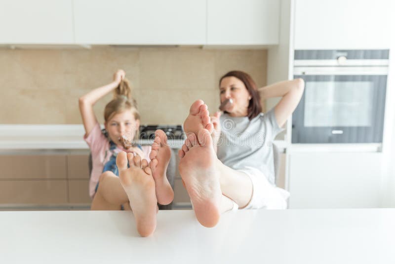 Cute girl and her mother are smiling while eating ice cream in the kitchen with feet on the table