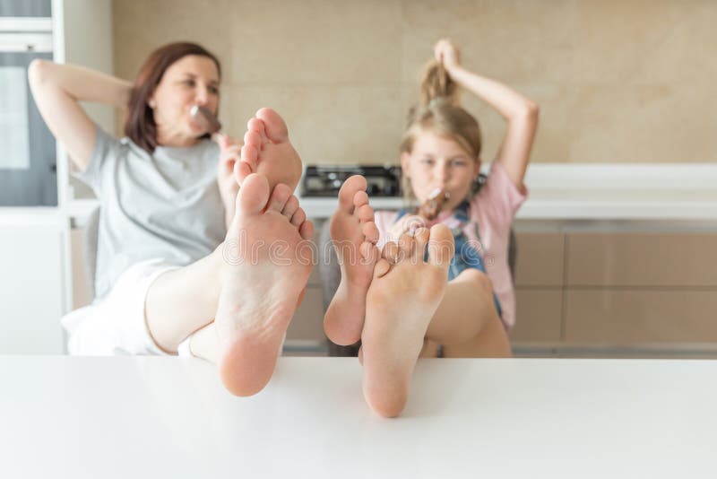 Cute girl and her mother are smiling while eating ice cream in the kitchen with feet on the table