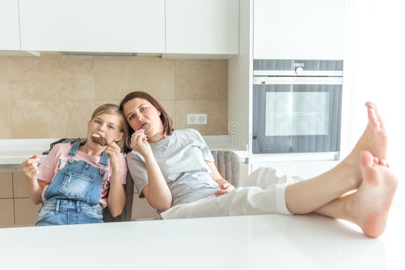 Cute girl and her mother are smiling while eating ice cream in the kitchen with feet on the table