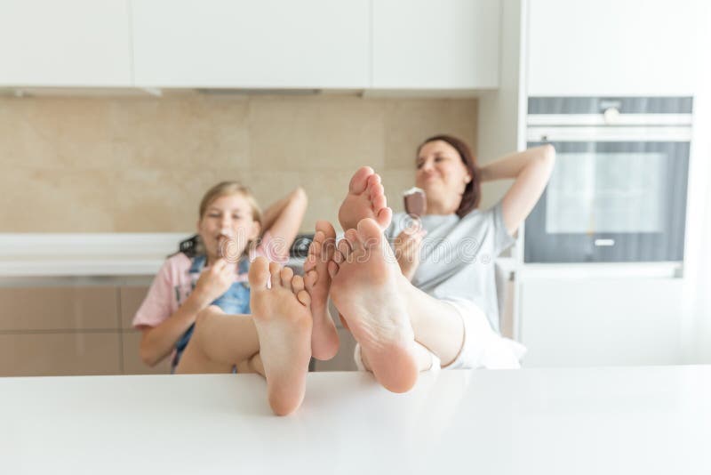 Cute girl and her mother are smiling while eating ice cream in the kitchen with feet on the table