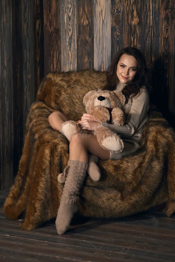 Cute girl in knee socks and sweater with Teddy bear in her hands sitting in armchair in wooden room