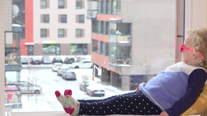 Cute girl with pink sunglasses sit on window sill and snowflakes fall outside