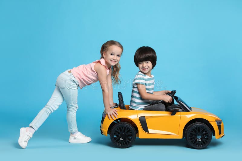 Cute girl pushing children`s electric toy car with little boy on light blue background