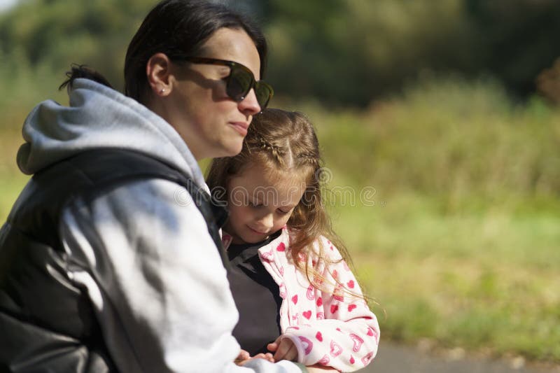 Cute Girl in Sunglasses vest and hoodie hugs hennes lilla dotter på en bänk. royaltyfri fotografi