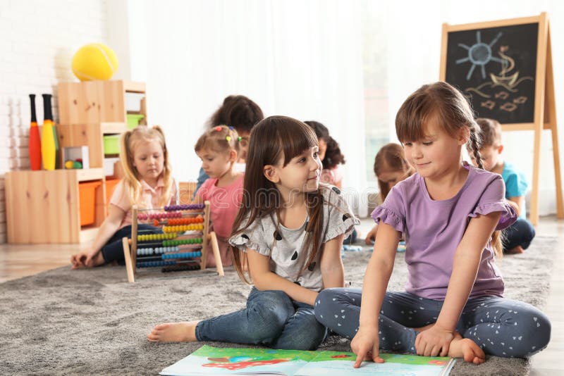 Cute Girls Reading Book on Floor while Other Children Playing Together ...