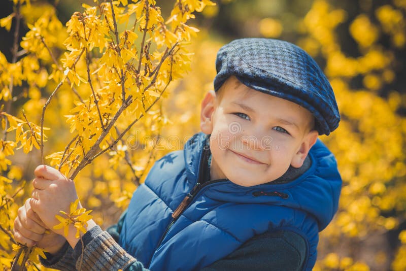 Cute handsome boy in stylish blue dress and hat close to yellow flowers enjoying spring time.