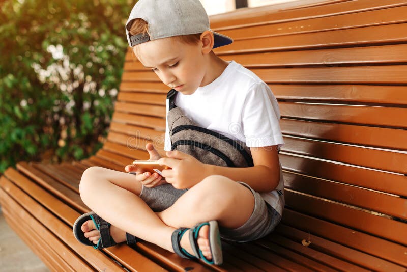 Cute kid playing games on smartphone outdoors. Leisure, children, technology and people. Boy plays online games on mobile phone. Kid sitting on bench at summer royalty free stock photos Cute kid playing games on smartphone outdoors. Leisure, children, technology and people. Boy plays online games on mobile phone. Kid sitting on bench at summer royalty free stock photos