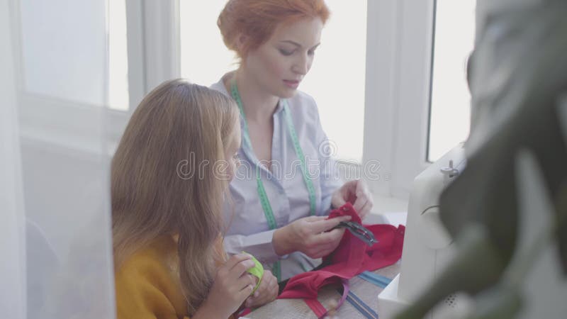 Cute Kleine Tochter untersucht Nähzubehör und lernt Nähen, während ihre Mutter mit rothahaariger Kleidung näht