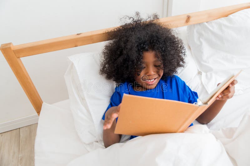 A cute little child African American girl reading a book while lying on the bed. Kid enjoy learning with happiness at home. Clever and smart learning concept