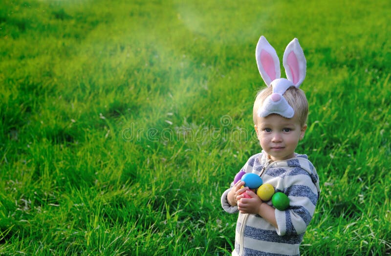 Cute little child on Easter day. Toddler boy with bunny ears hunts for Easter eggs on green lawn in nature or park. Easter egg