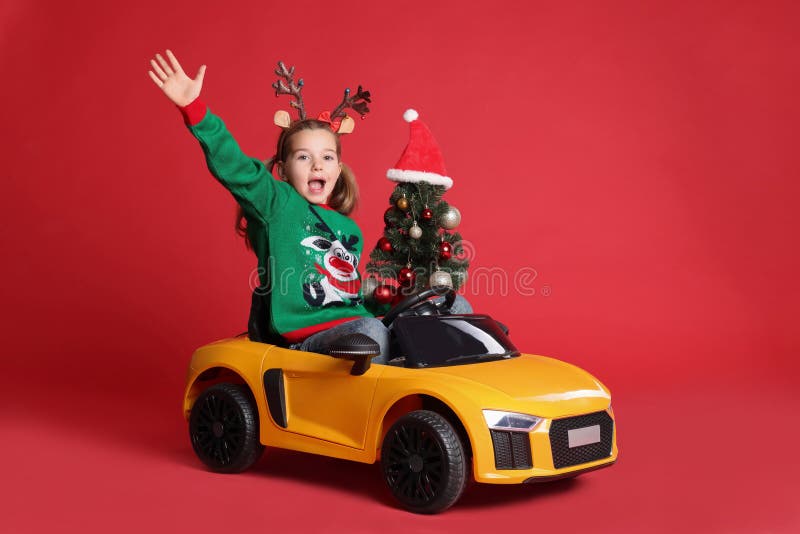 Cute little girl with Christmas tree driving children`s electric toy car on red background