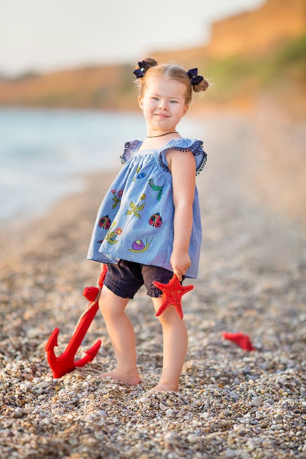 Cute little girl enjoying summer time on sea side beach happy playing with red star and tiny toy anchor on sand wearing nobby stock photography Cute little girl enjoying summer time on sea side beach happy playing with red star and tiny toy anchor on sand wearing nobby stock photography