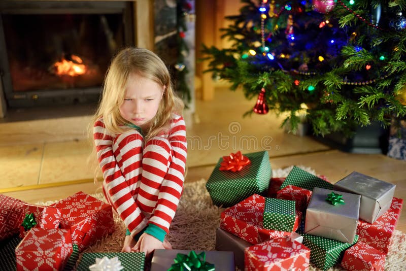 Cute little girl feeling unhappy with her Christmas gifts. Child sitting by a fireplace in a cozy dark living room on Xmas eve.