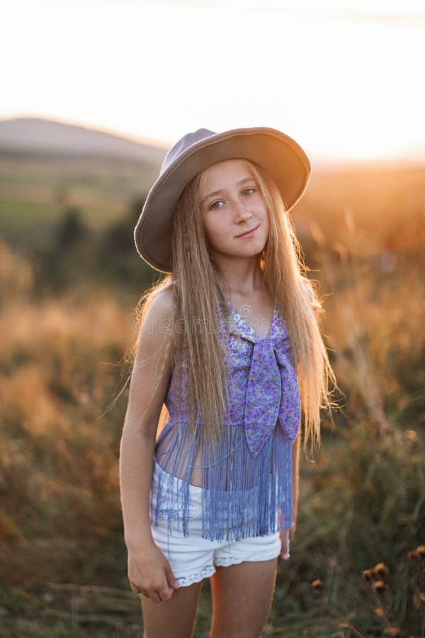 Cute little girl with long blond hair, wearing brown hat, posing in the wild flower and grass field with sunset