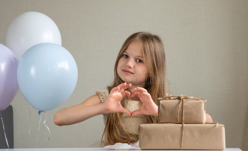 Cute little girl making heart with her hands and smiling surrounded by air balloons and gifts. Adorable kid smiling in royalty free stock photos