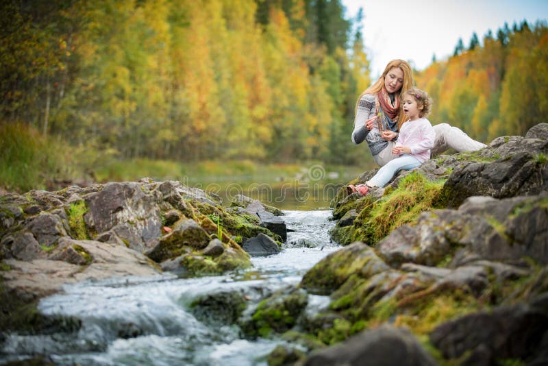 Cute little girl and mother sitting on a rock in autumn forest at stream