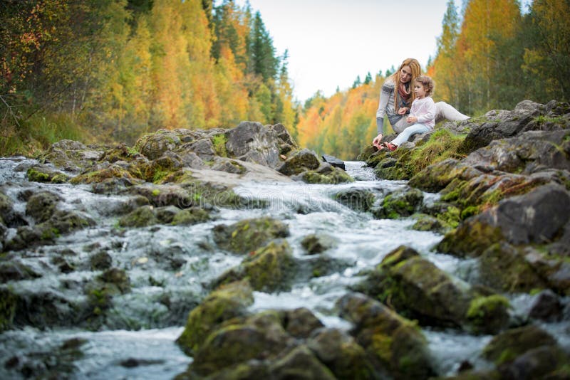 Cute little girl and mother sitting on a rock in autumn forest at stream