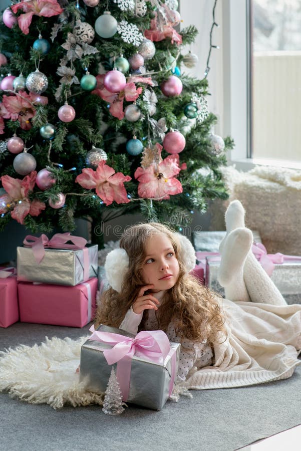 Cute little girl in winter clothes posing in home front of Christmas tree with gift box in hand