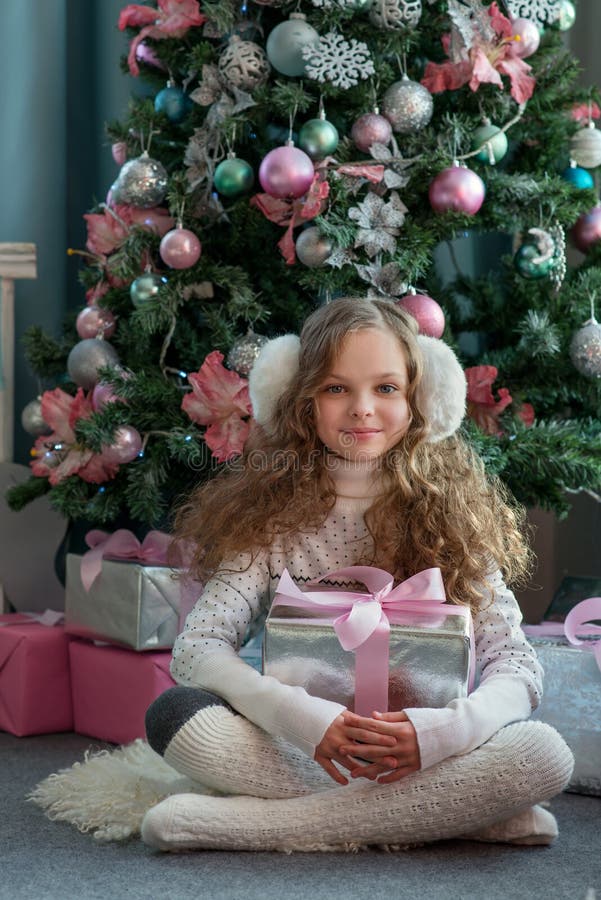 Cute little girl in winter clothes posing in home front of Christmas tree with gift box in hand