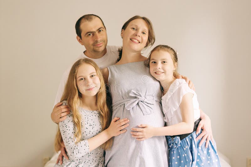 Cute girls and her handsome dad are hugging her beautiful pregnant mom`s tummy, looking at camera and smiling.