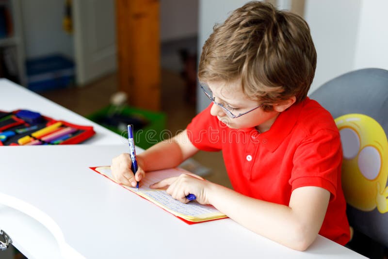 Cute little kid boy with glasses at home making homework, writing letters with colorful pens.