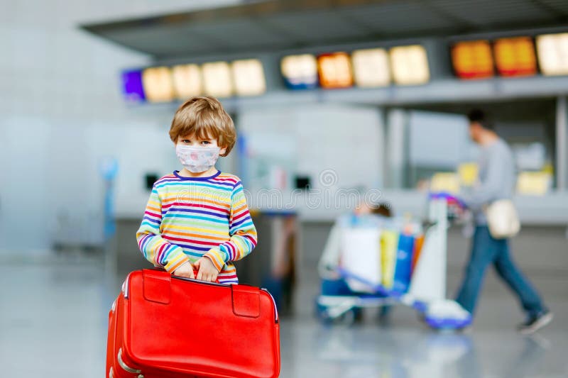 Cute little kid boy with medical mask and luggage, red suitcase on international airport. Family travel during corona. Virus and covid pandemic