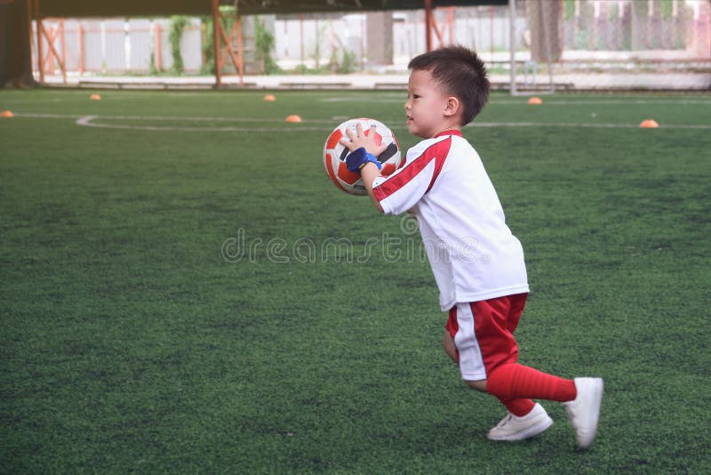 Cute little smiling Asian kindergarten kid, football player in soccer uniform is playing football at Training Session, Soccer