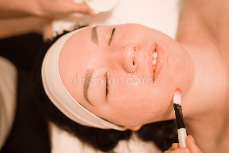 Cute mix race woman lies and enjoys the process of applying a brush mask in the beauty salon on her face stock photo Cute mix race woman lies and enjoys the process of applying a brush mask in the beauty salon on her face stock photo