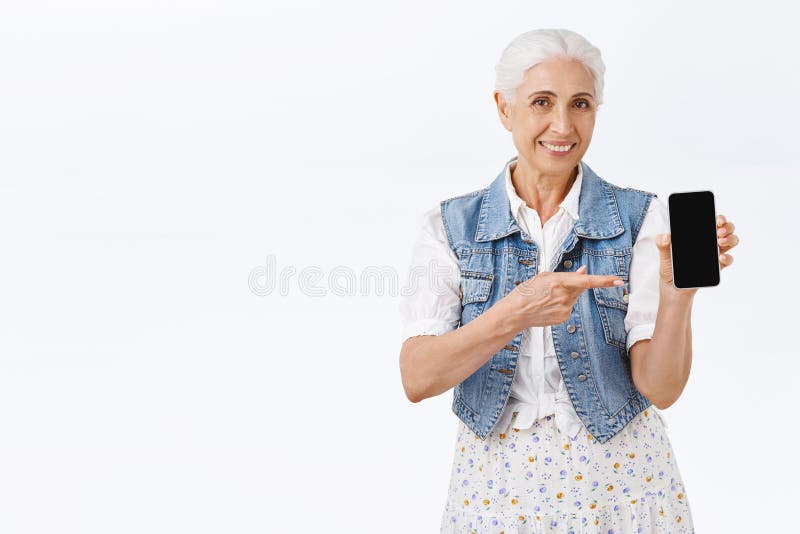 Cute modern old lady with combed grey hair showing smartphone display, pointing at mobile screen and smiling as royalty free stock images Cute modern old lady with combed grey hair showing smartphone display, pointing at mobile screen and smiling as royalty free stock images