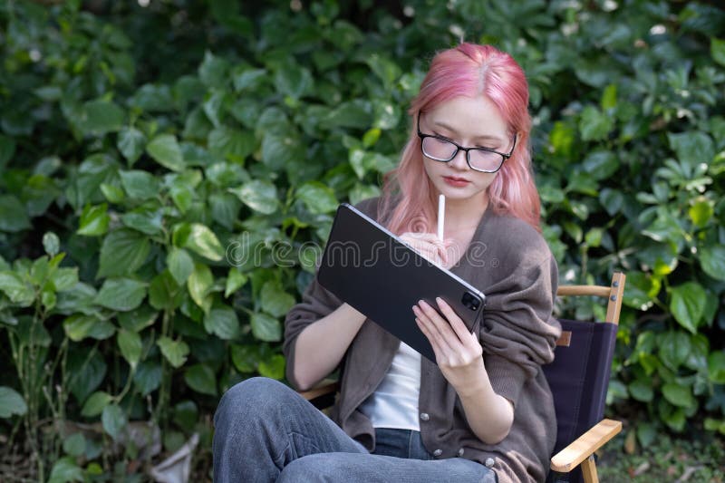 Cute and positive Asian woman recording data on a tablet or jotting down some ideas while sitting on a chair under a