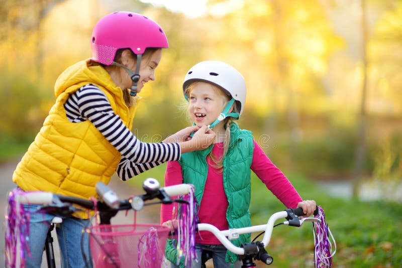 Cute sisters riding bikes in a city park on sunny autumn day. Active family leisure with kids. Children wearing safety hemet while