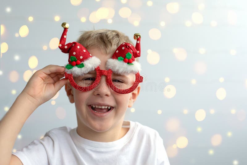 Cute smiling boy with blonde hair in festive Christmas glasses on a light background with bokeh